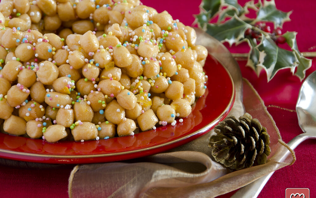 Struffoli campani coperti di miele e confettini colorati su un piatto rosso, con agrifoglio e pigna: la stessa forma dei purceddhruzzi salentini