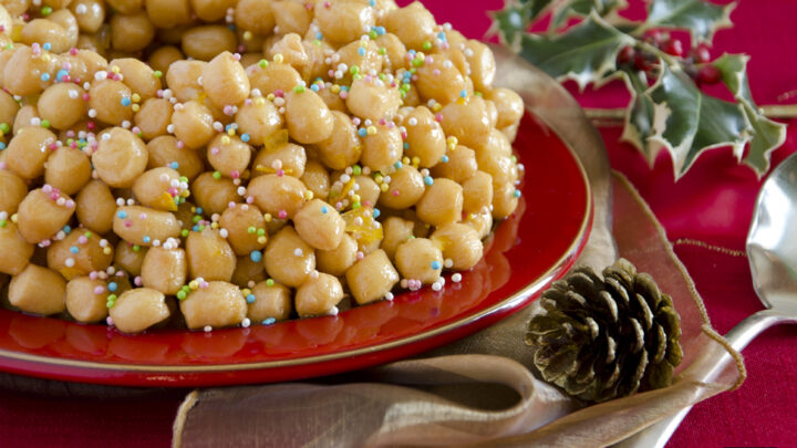 Struffoli campani coperti di miele e confettini colorati su un piatto rosso, con agrifoglio e pigna: la stessa forma dei purceddhruzzi salentini